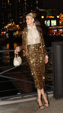 By the waterfront at night, a person wears Social Queen’s Sequin Velvet Skirt with a matching jacket and sheer white blouse, holding a sparkly handbag. City lights and buildings shimmer in the background.
