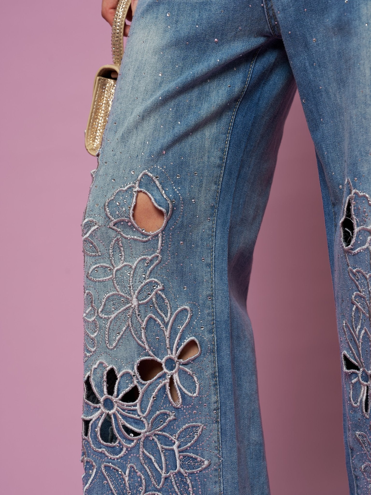 Close-up of Social Queen Cutout Embroidered Rhinestone Jeans, featuring wide legs in washed denim with floral cutout embroidery and rhinestone details, styled with a gold handbag against a pink backdrop.