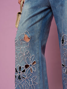 Close-up of Social Queen Cutout Embroidered Rhinestone Jeans, featuring wide legs in washed denim with floral cutout embroidery and rhinestone details, styled with a gold handbag against a pink backdrop.