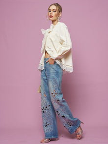 A woman stands against a pink background wearing a white blouse, silver high-heeled sandals, and Social Queen Cutout Embroidered Rhinestone Jeans.