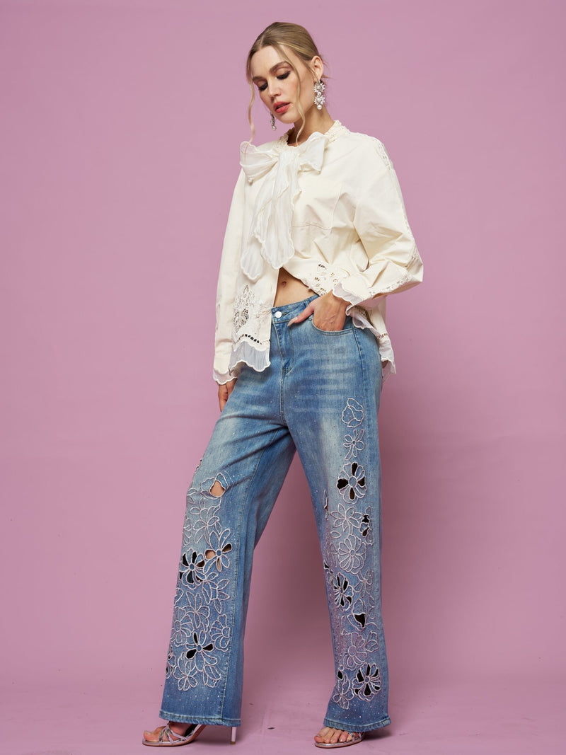 A person stands against a pink background wearing a white ruffled blouse and Social Queen Cutout Embroidered Rhinestone Jeans, styled with clear heeled sandals.