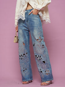 A person wears Social Queen Cutout Embroidered Rhinestone Jeans and a cream blouse with strappy heels, holding a small gold handbag against a pink background.