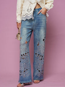 A model wears Social Queen’s Cutout Embroidered Rhinestone Jeans with a cream blouse, holding a small gold handbag, set against a pink background.