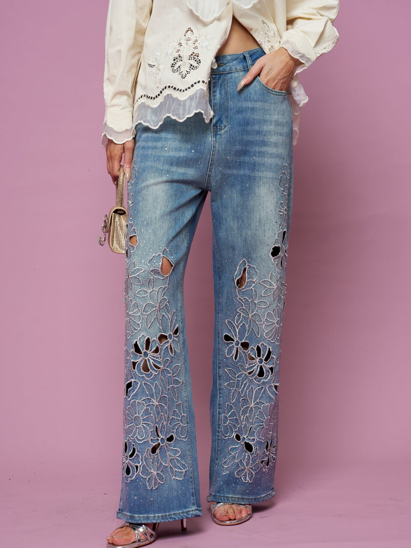 A model wears Social Queen’s Cutout Embroidered Rhinestone Jeans with a cream blouse, holding a small gold handbag, set against a pink background.