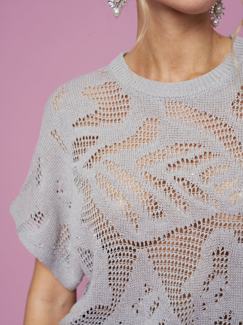 A model wears the Social Queen Cutout Jacquard Knit Top in light gray, featuring botanical cutouts, against a pink background. Only the lower face and upper body are shown.