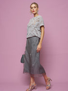 A woman stands against a pink background, wearing the Social Queen Cutout Jacquard Knit Top, a metallic gray midi skirt, silver heels, statement earrings, and holding a small purse.
