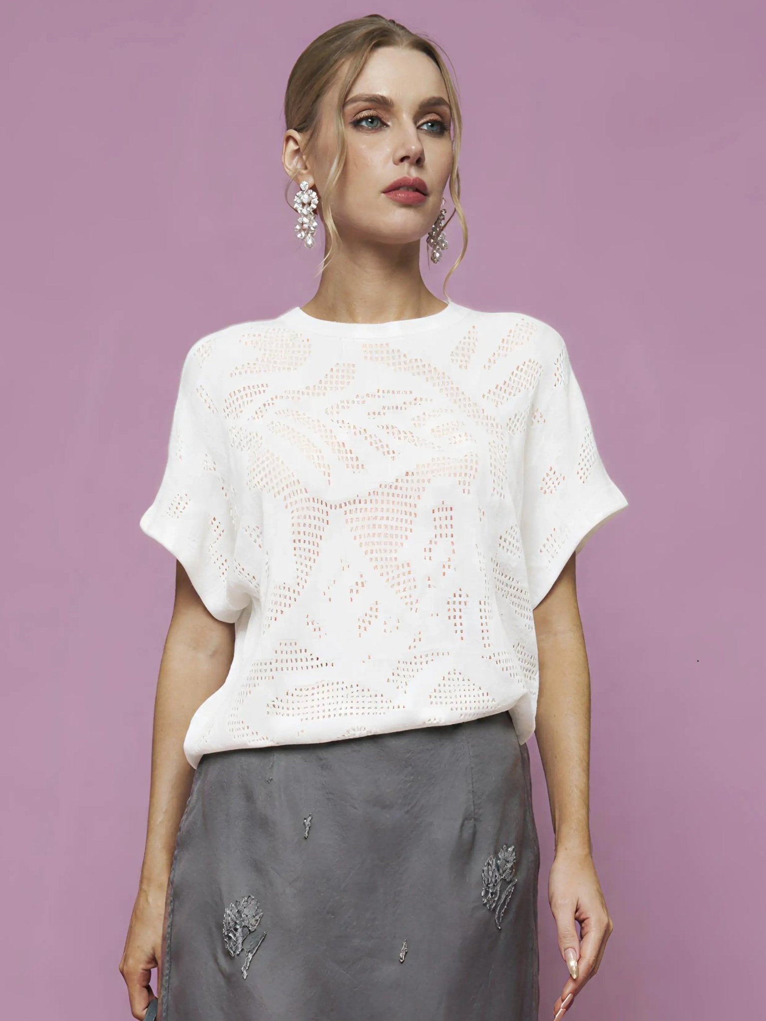 A woman stands against a pink background, wearing the Social Queen Cutout Jacquard Knit Top in white, paired with a gray embroidered skirt and large dangling earrings.