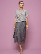 A woman stands against a pink background wearing the Social Queen Cutout Jacquard Knit Top, a sheer grey midi skirt, silver sandals, statement earrings, and holding a small handbag.