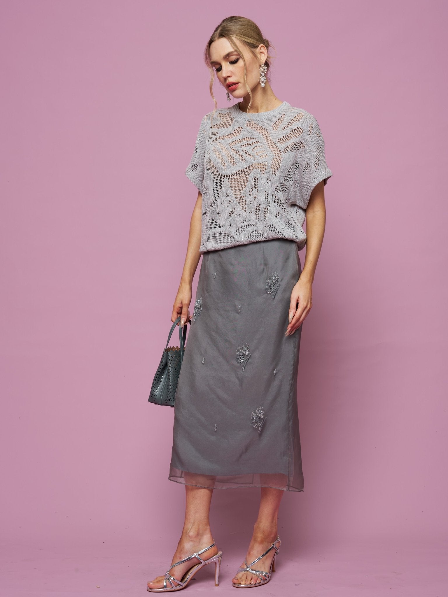 A woman poses against a pink background wearing the Social Queen Cutout Jacquard Knit Top in light gray, paired with a gray midi skirt, silver heels, and carrying a small dark handbag.