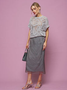 A woman poses against a pink background wearing the Social Queen Cutout Jacquard Knit Top in light gray, paired with a gray midi skirt, silver heels, and carrying a small dark handbag.