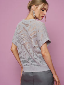Woman with blonde hair in an updo wears dangly earrings, a Social Queen Cutout Jacquard Knit Top in light purple, and a dark skirt, standing against a pink background.
