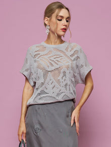 A woman stands against a pink background wearing the Social Queen Cutout Jacquard Knit Top with batwing sleeves and a grey skirt, holding a handbag and looking down to her left.