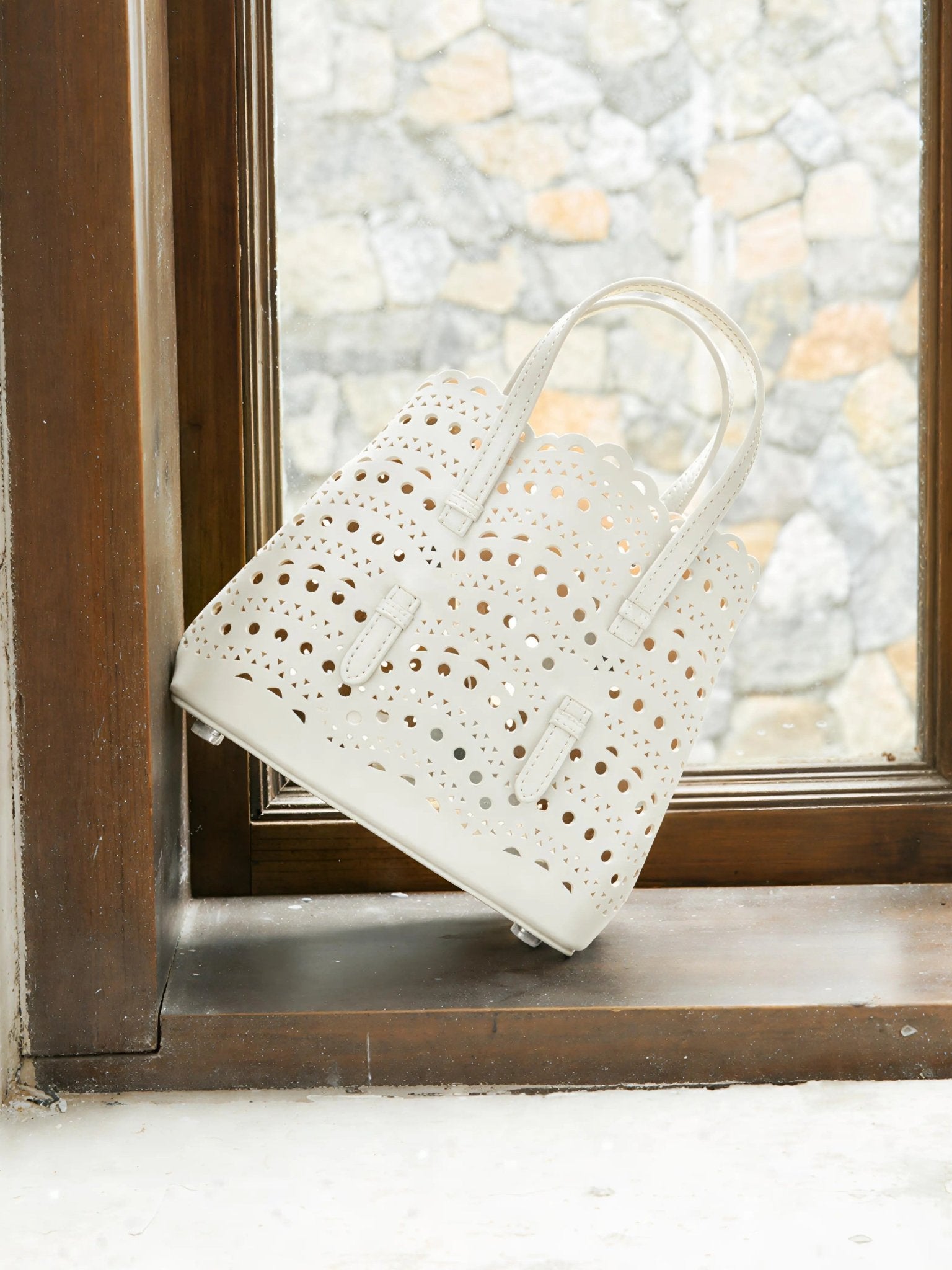 The Social Queen Cutout Leather Top-Handle Bag, crafted in white genuine leather with scalloped edges and a geometric cutout design, is placed on a window sill overlooking a stone wall.