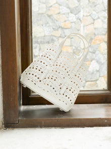 The Social Queen Cutout Leather Top-Handle Bag, crafted in white genuine leather with scalloped edges and a geometric cutout design, is placed on a window sill overlooking a stone wall.