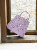 The Social Queen Cutout Leather Top-Handle Bag in lavender, featuring scalloped edges and a geometric cutout design, rests on a windowsill against a stone wall backdrop.