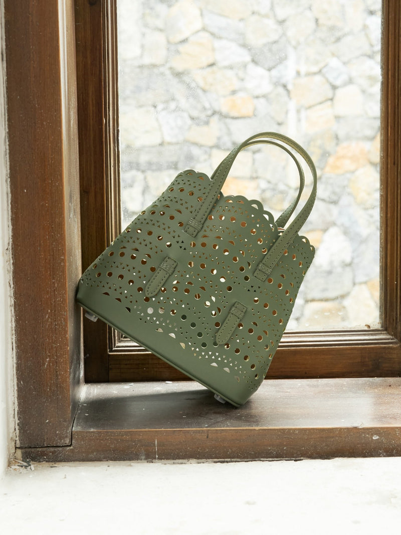 The Social Queen Cutout Leather Top-Handle Bag in olive green rests against a wooden window frame, showcasing its geometric cutout design with a stone wall visible outside.