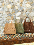 Three Social Queen Cutout Leather Top-Handle Bags in beige, green, and brown are displayed on a patterned bench with a stone wall behind them. Each mini bag features a chic geometric cutout design.