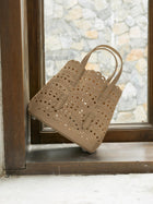 The Social Queen Cutout Leather Top-Handle Bag, featuring a tan geometric cutout design and double handles, rests on a wooden window ledge with a stone wall visible outside.