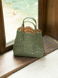 The Social Queen Cutout Leather Top-Handle Bag, made from green genuine leather with scalloped edges and geometric cutouts, sits on a wooden windowsill beside a glass window.