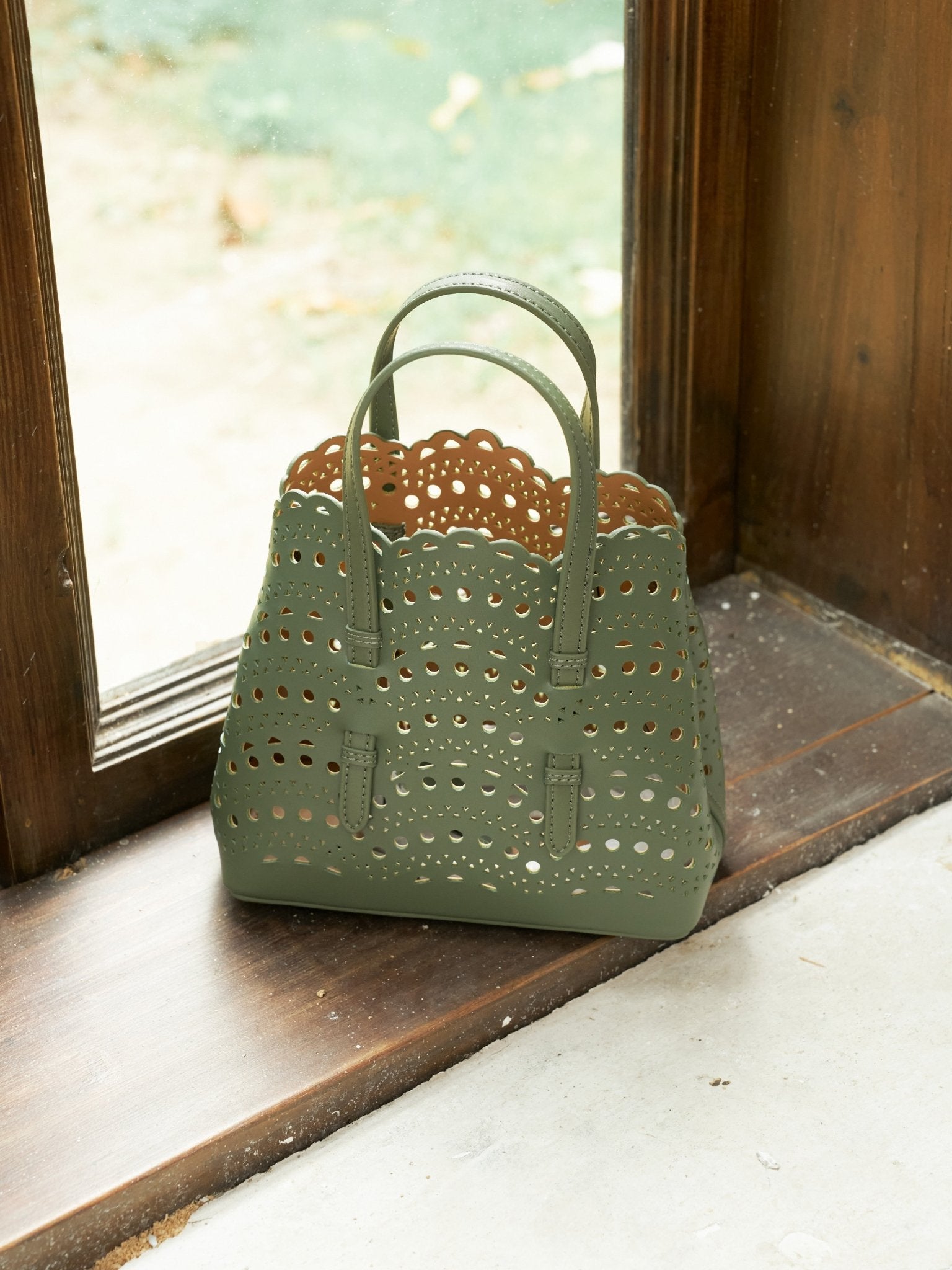 The Social Queen Cutout Leather Top-Handle Bag, made from green genuine leather with scalloped edges and geometric cutouts, sits on a wooden windowsill beside a glass window.