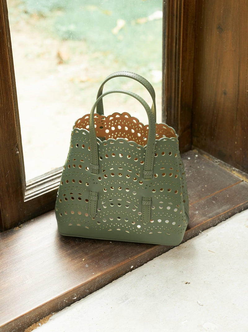 The Social Queen Cutout Leather Top-Handle Bag, made from green genuine leather with scalloped edges and geometric cutouts, sits on a wooden windowsill beside a glass window.