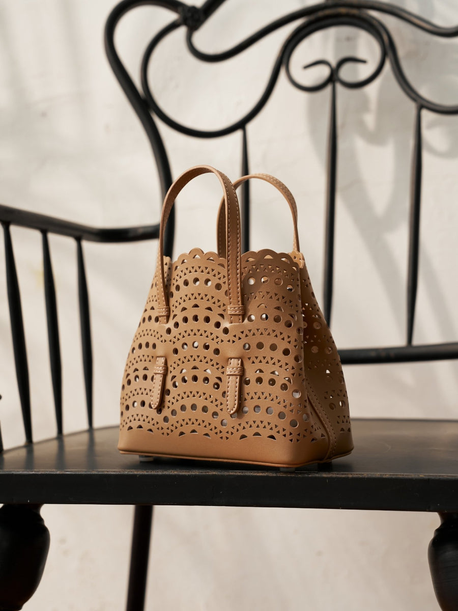 The Cutout Leather Top-Handle Mini Bag by Social Queen, featuring geometric cutouts and scalloped edges in tan genuine leather, is displayed on a black wrought-iron chair.