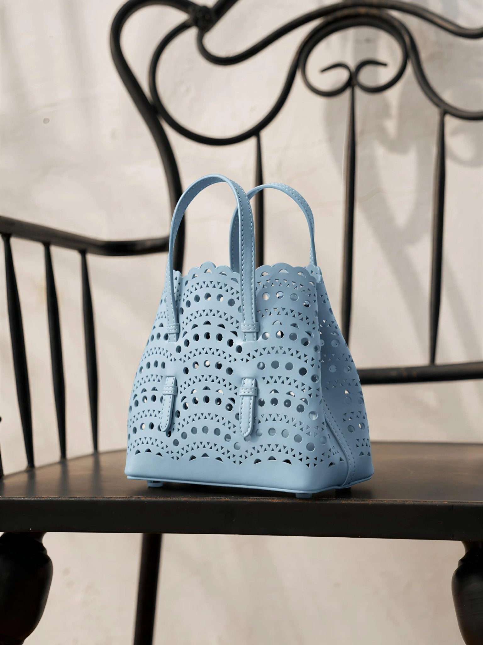 The Social Queen Cutout Leather Top-Handle Mini Bag, in light blue with a geometric cutout design and two handles, rests on a dark wooden chair featuring a decorative metal backrest.