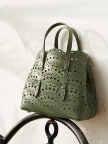 The Social Queen Cutout Leather Top-Handle Mini Bag, crafted in olive green genuine leather with double handles and a geometric cutout design, rests against a white wall.