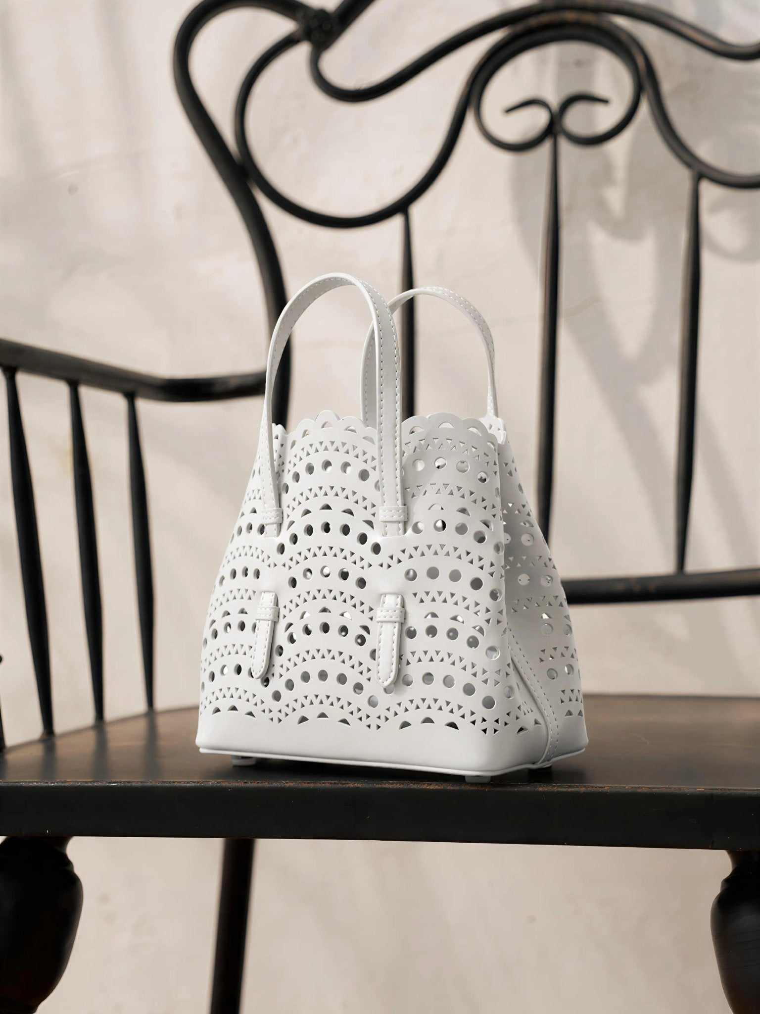 The Social Queen Cutout Leather Top-Handle Mini Bag, featuring a white geometric design, sits on a black wooden chair with a decorative metal backrest.