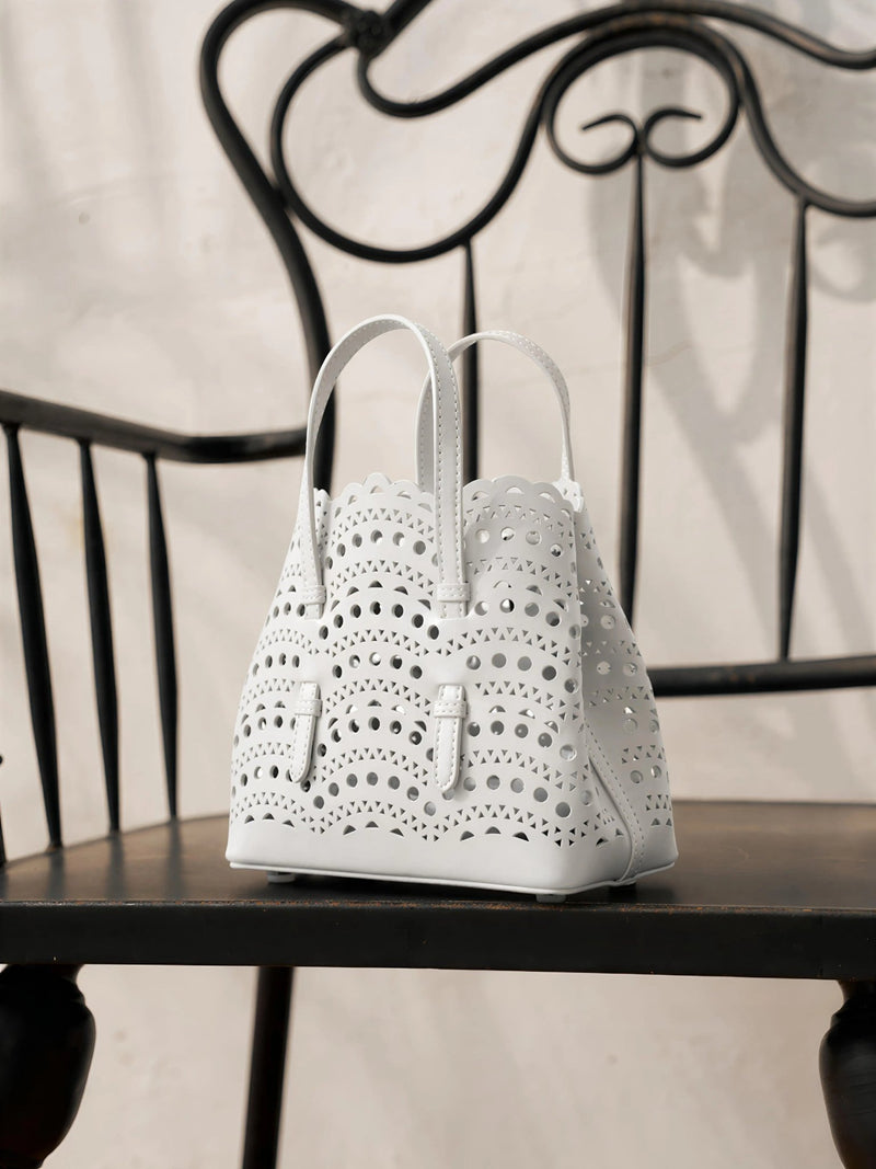 The Social Queen Cutout Leather Top-Handle Mini Bag, featuring a white geometric design, sits on a black wooden chair with a decorative metal backrest.