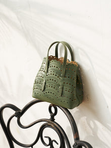 The Social Queen Cutout Leather Top-Handle Mini Bag, in green genuine leather with a geometric cutout design and scalloped edges, is displayed on a black metal chair against a white wall.