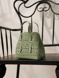 The Social Queen Cutout Leather Top-Handle Mini Bag, in light green with scalloped edges and a geometric cutout design, rests on a black wooden chair with a decorative metal back.