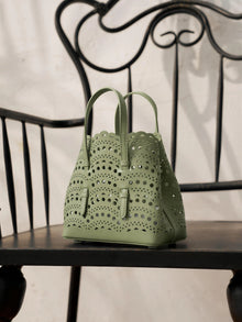The Social Queen Cutout Leather Top-Handle Mini Bag, in light green with scalloped edges and a geometric cutout design, rests on a black wooden chair with a decorative metal back.