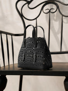 The Social Queen Cutout Leather Top-Handle Mini Bag, featuring a black scalloped geometric design, rests on a black metal chair with curved and straight lines.