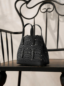 The Social Queen Cutout Leather Top-Handle Mini Bag, featuring a black scalloped geometric design, rests on a black metal chair with curved and straight lines.