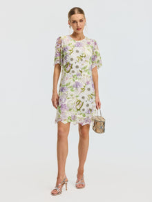 A woman poses against a plain background in the Social Queen Dimensional Embroidered Beaded Lace Dress, knee-length with short sleeves, styled with a small woven handbag and strappy heeled sandals.