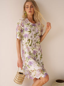 A woman models the Social Queen Dimensional Embroidered Beaded Lace Dress in purple, green, and white while holding a beaded handbag against a plain wall.