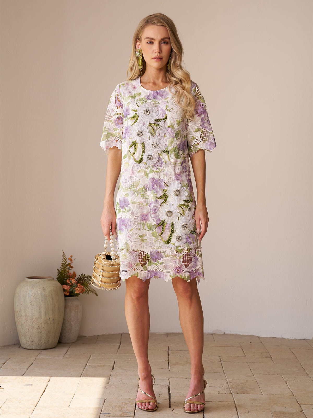 A woman stands indoors wearing the Social Queen Dimensional Embroidered Beaded Lace Dress in white, green, and purple, styled with a small woven handbag and heeled sandals.