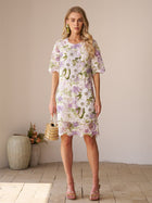 A woman stands indoors wearing the Social Queen Dimensional Embroidered Beaded Lace Dress in white, green, and purple, styled with a small woven handbag and heeled sandals.