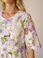A woman wears the Social Queen Dimensional Embroidered Beaded Lace Dress, featuring white lace, green and purple dimensional embroidery, beaded brooches, short sleeves, and intricate cutouts for a stunningly elegant look.