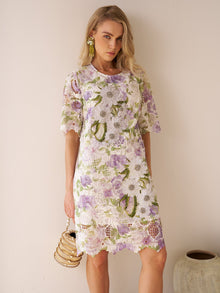 A woman models the Social Queen Dimensional Embroidered Beaded Lace Dress—a purple and green knee-length design—while holding a beaded handbag beside a ceramic vase against a plain light-colored wall.