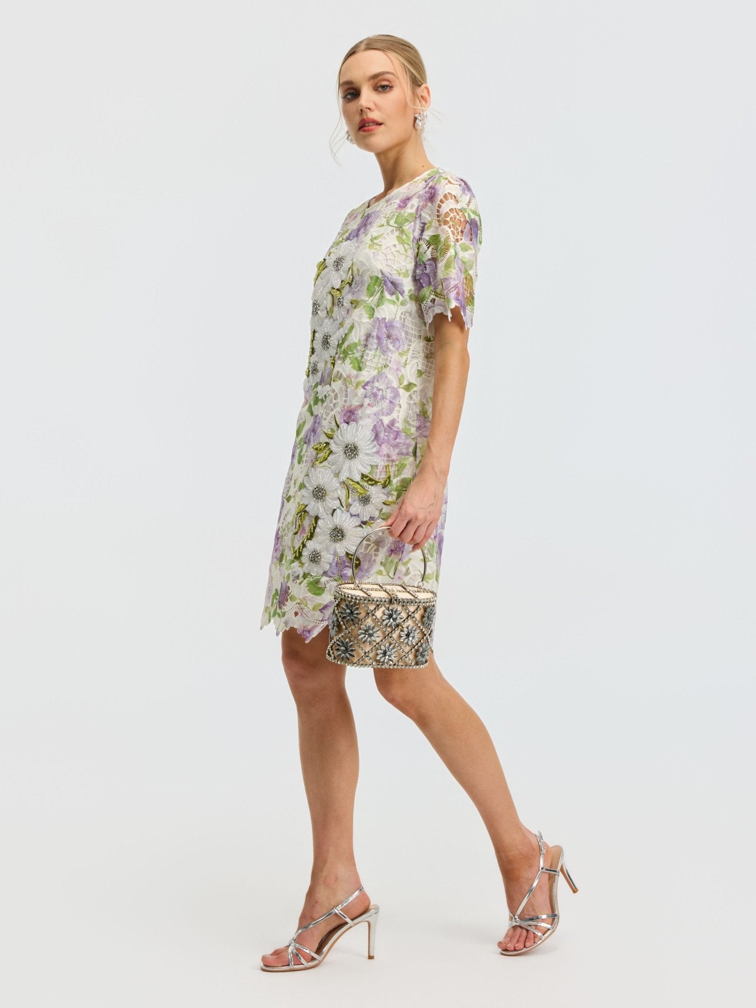 A woman wears the Social Queen Dimensional Embroidered Beaded Lace Dress with silver heels, holding a small patterned handbag, and poses against a plain white background.