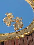 The Dimensional Floral Earrings by Social Queen rest on an ornate gold mirror atop wood, reflecting the blue sky—a stunning, elegant accessory for any wedding guest.