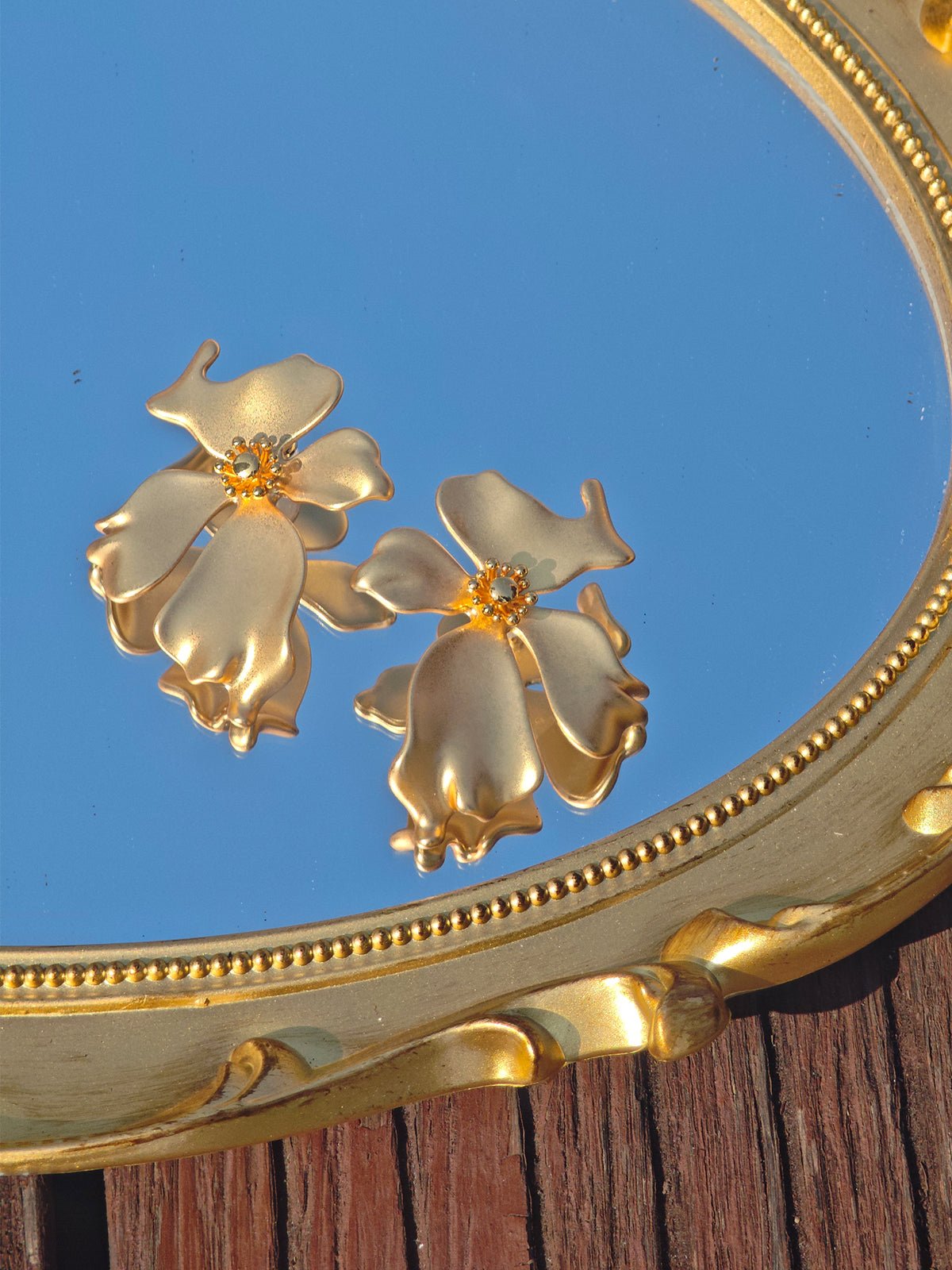 The Dimensional Floral Earrings by Social Queen rest on an ornate gold mirror atop wood, reflecting the blue sky—a stunning, elegant accessory for any wedding guest.