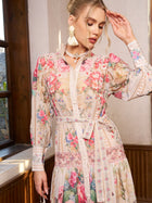 A woman in the Social Queen Eden Garden Print Tie-Waist Dress stands indoors by a window, touching her forehead with her hand.