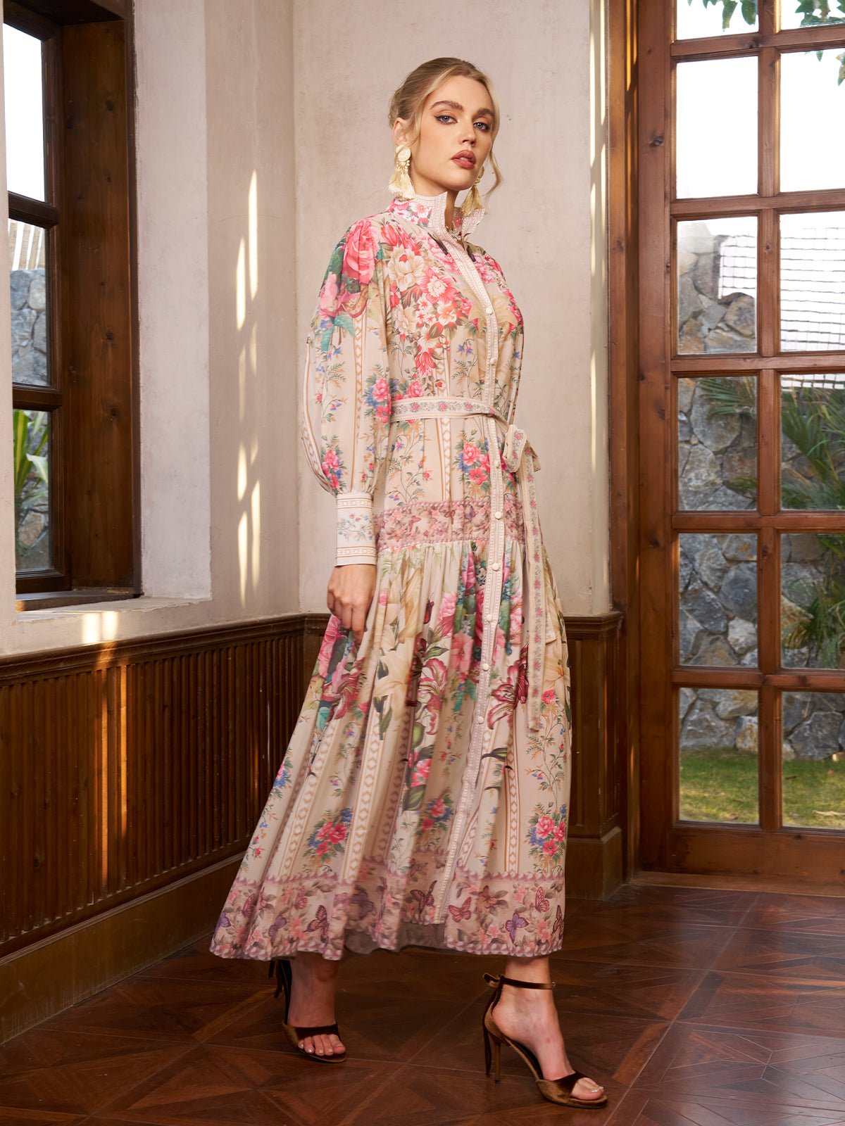 A woman stands indoors in the Social Queen Eden Garden Print Tie-Waist Dress, featuring a silk-look floral pattern, long sleeves, lace details, and a high collar. She pairs it with high-heeled sandals as sunlight streams through wooden framed windows.