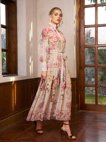A woman stands indoors in the Social Queen Eden Garden Print Tie-Waist Dress, featuring a silk-look floral pattern, long sleeves, lace details, and a high collar. She pairs it with high-heeled sandals as sunlight streams through wooden framed windows.