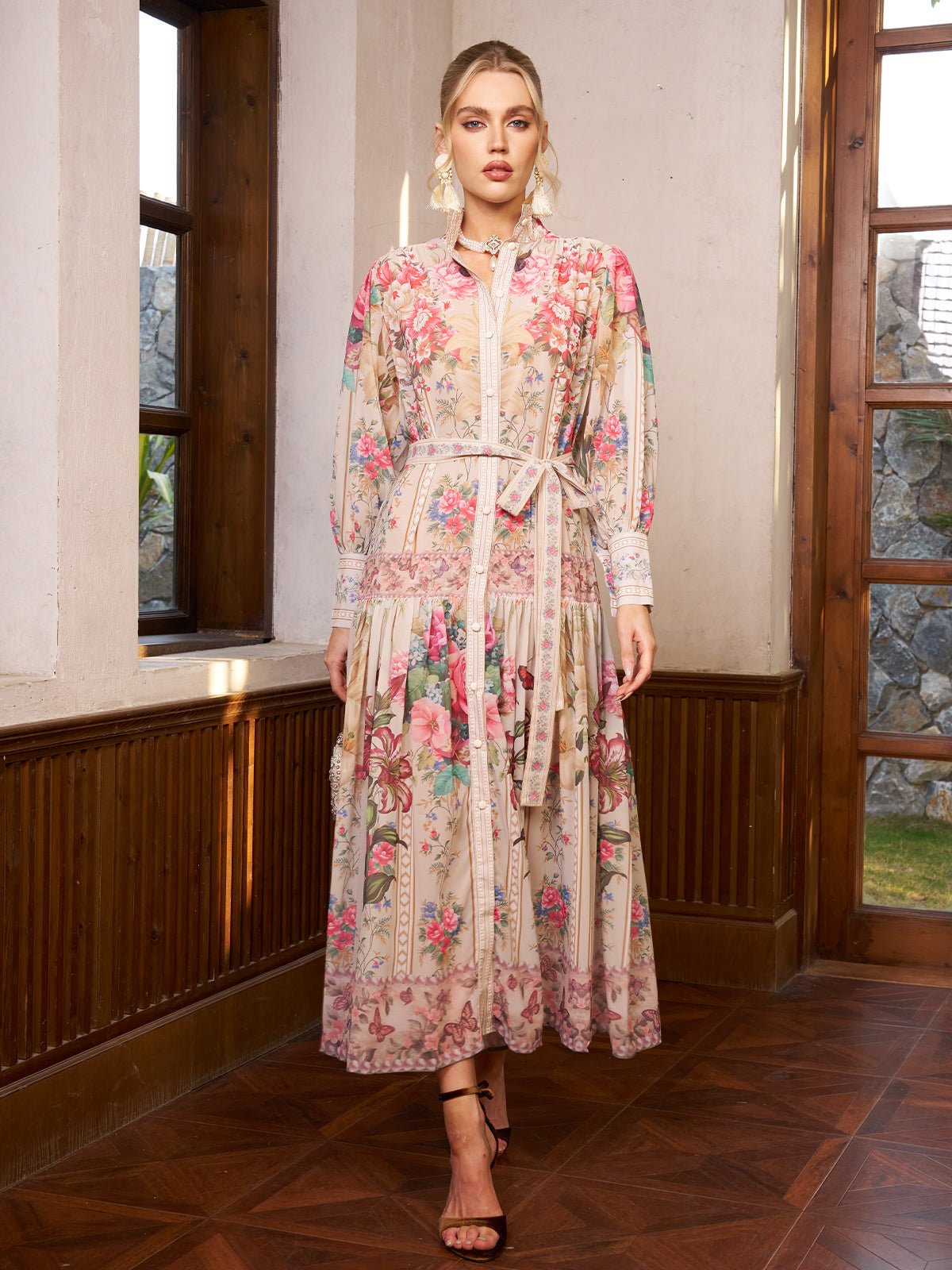 A woman stands indoors wearing the Social Queen Eden Garden Print Tie-Waist Dress with sheer sleeves, styled with strappy heels.