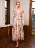 A woman stands indoors wearing the Social Queen Eden Garden Print Tie-Waist Dress with sheer sleeves, styled with strappy heels.