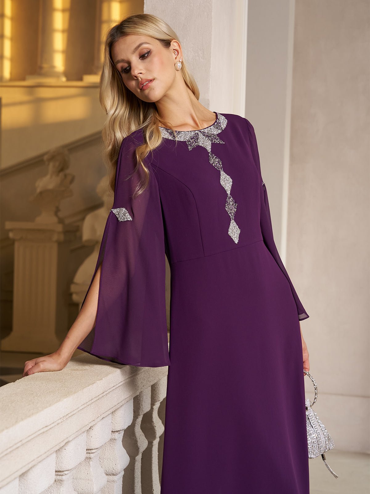 A woman stands indoors by a white stone railing, holding a silver handbag and wearing the Social Queen Elegant Beaded Split-Sleeve Dress—a long-sleeve purple chiffon cocktail dress with silver embellishments.